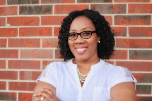 Black woman wearing a white shirt and glasses, standing in front of a brick wall with arms folded.