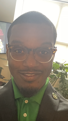 Black man wearing glasses, green button up shirt, and a black jacket.